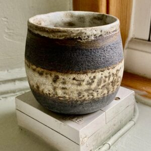 Artisan functional pottery — small rustic tea cup made from black clay with cream and dark glaze bands, sitting on a white surface near a window frame.